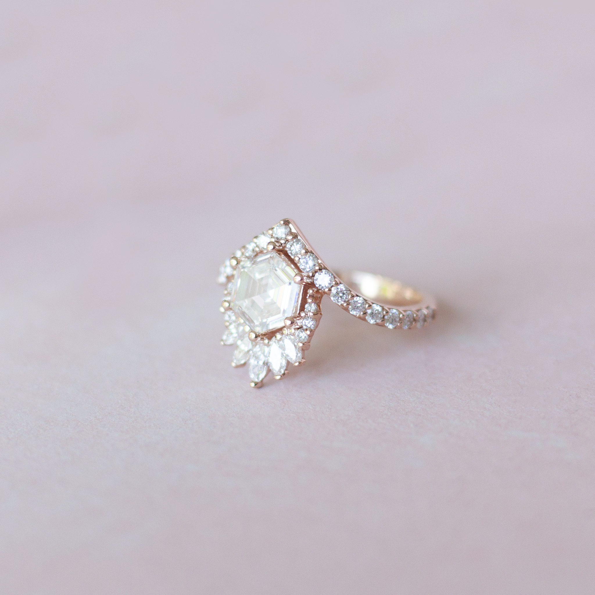 Unique step cut hexagon diamond engagement ring, featuring a delicate V shape band, encrusted with round brilliant diamonds. The hexagon step cut diamond has a low profile setting and is surrounded with a fine crown of marquise and round cut white diamonds. Made in 14k or 18k rose gold.